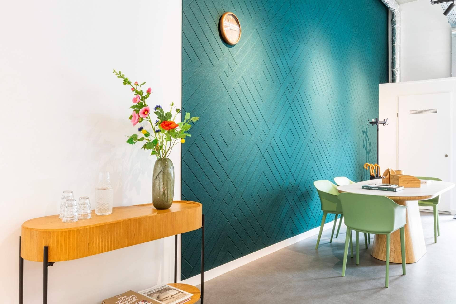 Sfeerbeeld van een betonnen wand met diepgroen akoestisch wandpaneel, met een V-patroon. Naast het paneel zien we een stijlvol houten tafeltje met decoratieve vaas.