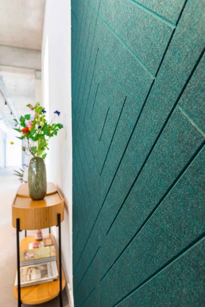 Close-up van een betonnen wand met diepgroen akoestisch wandpaneel, met een V-patroon. Naast het paneel zien we een stijlvol houten tafeltje met decoratieve vaas.
