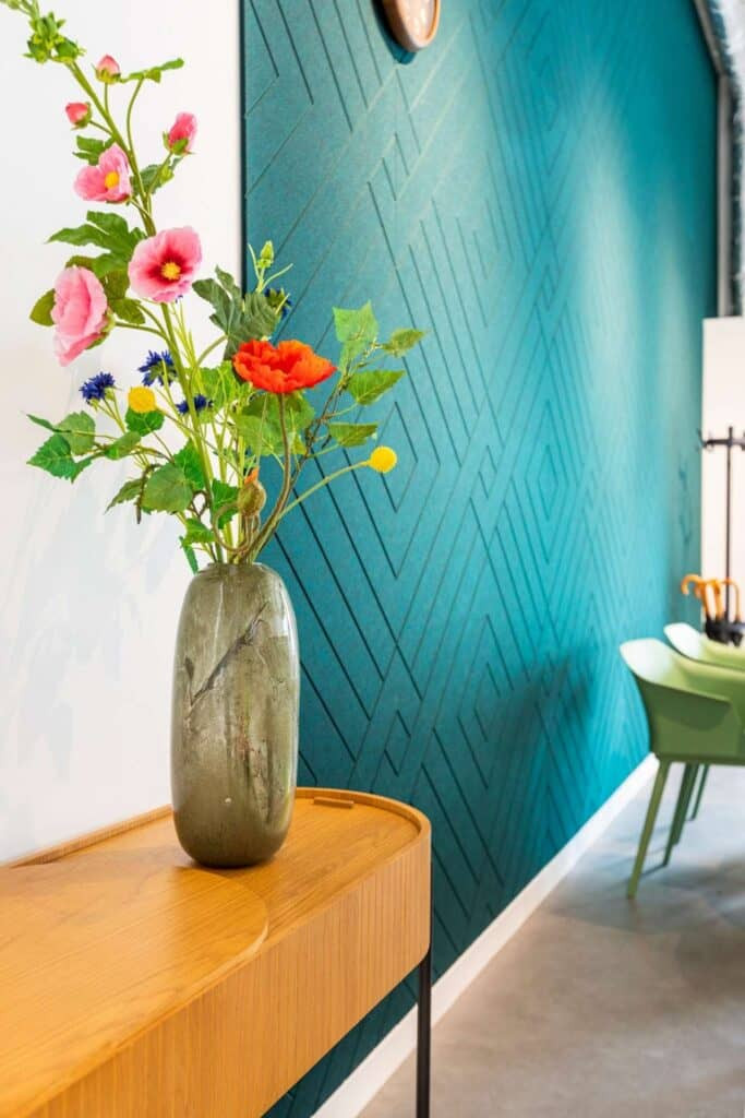 Sfeerbeeld van een betonnen wand met blauw akoestisch wandpaneel, met een V-patroon. Naast het paneel zien we een stijlvol houten tafeltje met decoratieve vaas.