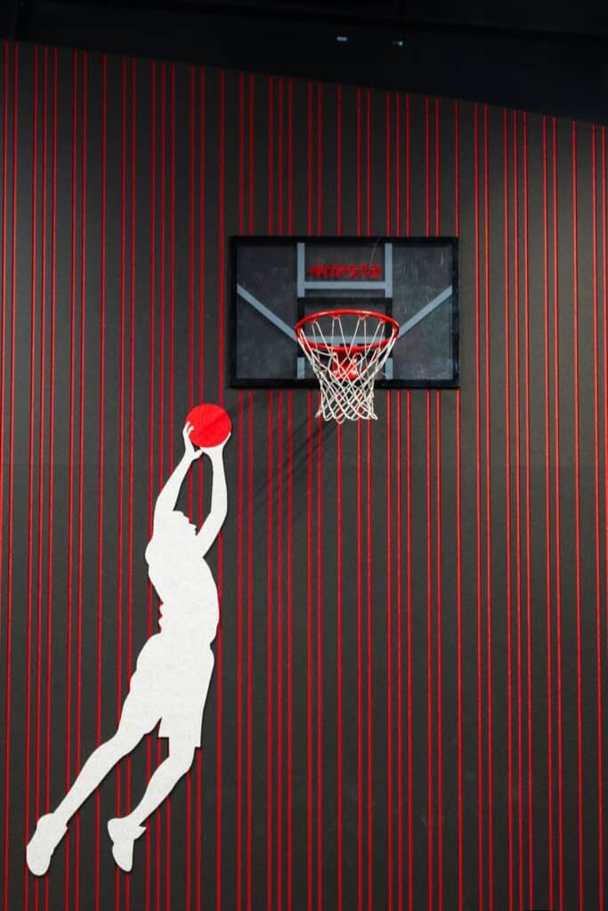 Impressie van een sportruimte met basket aan een lattenwand van akoestisch PET-vilt. Ook zien we een PET-vilt uitsnede van een sportend figuur aan de wand, die een slam dunk maakt.